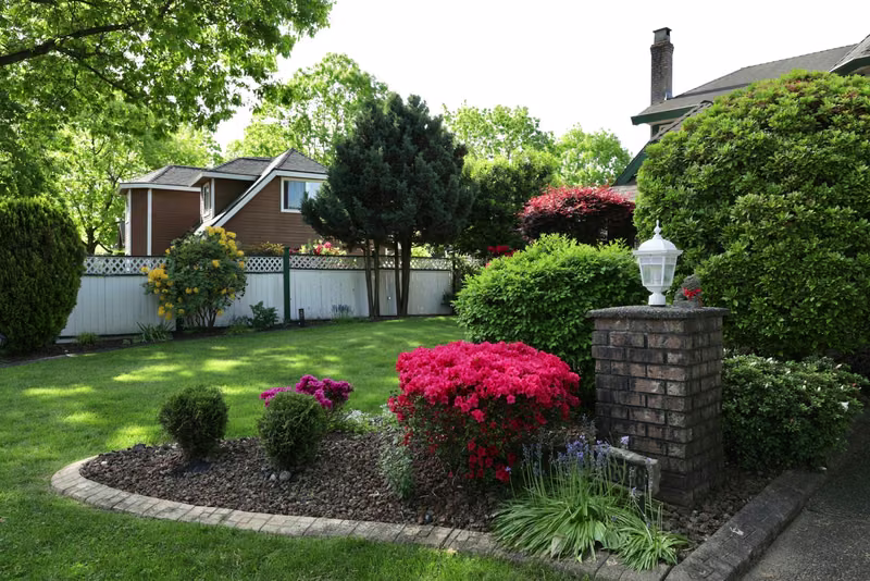 Community entrance garden with red azaleas, fresh mulch beds, a brick pillar, and a manicured lawn