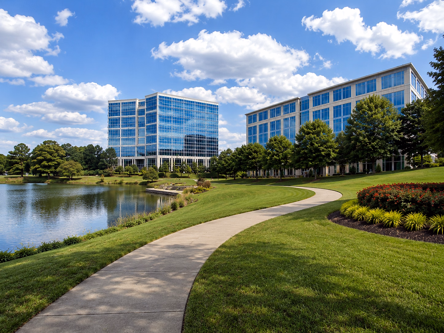 A manicured lawn and curved walkway leading to a modern glass office campus beside a lake